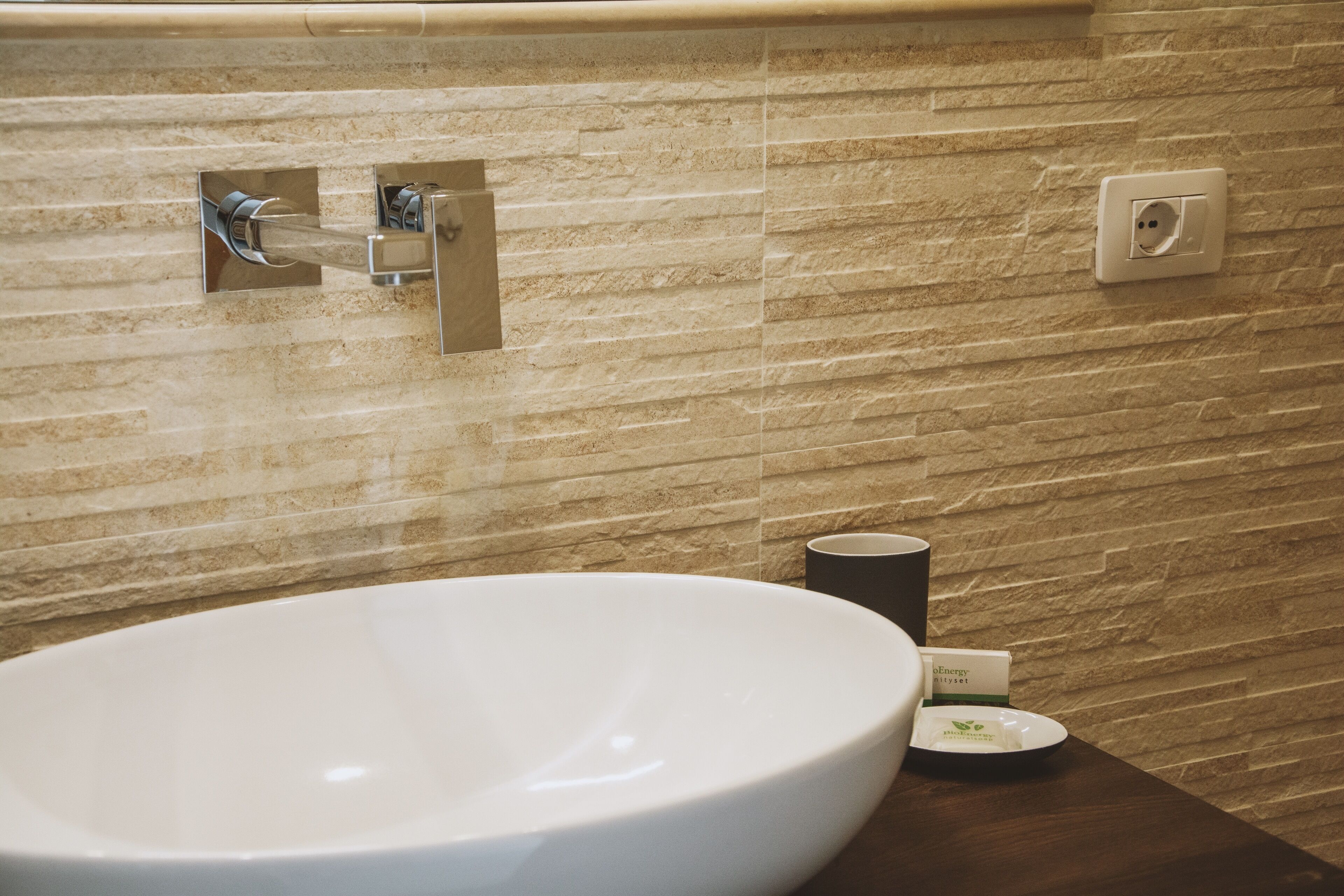 A bathroom with a large white sink, a towel rack, a power outlet, and a small dish with a green logo.