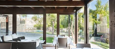 Un área de comedor al aire libre con mobiliario de mimbre y mesas, amplias ventanas con vistas a un jardín y una piscina.