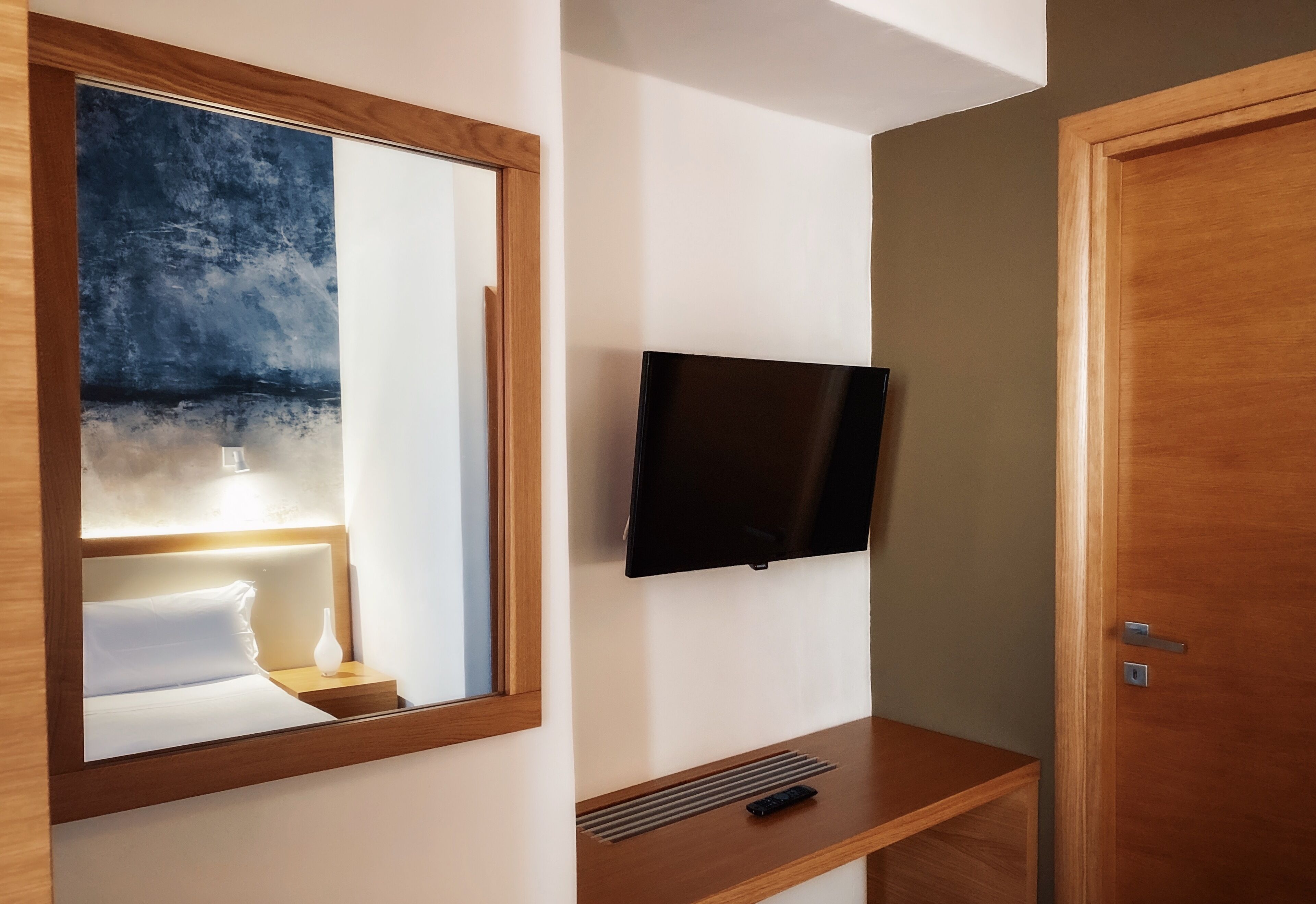 A hotel room with a bed, a bedside table, a mirror, a television mounted on the wall, and a wooden door.