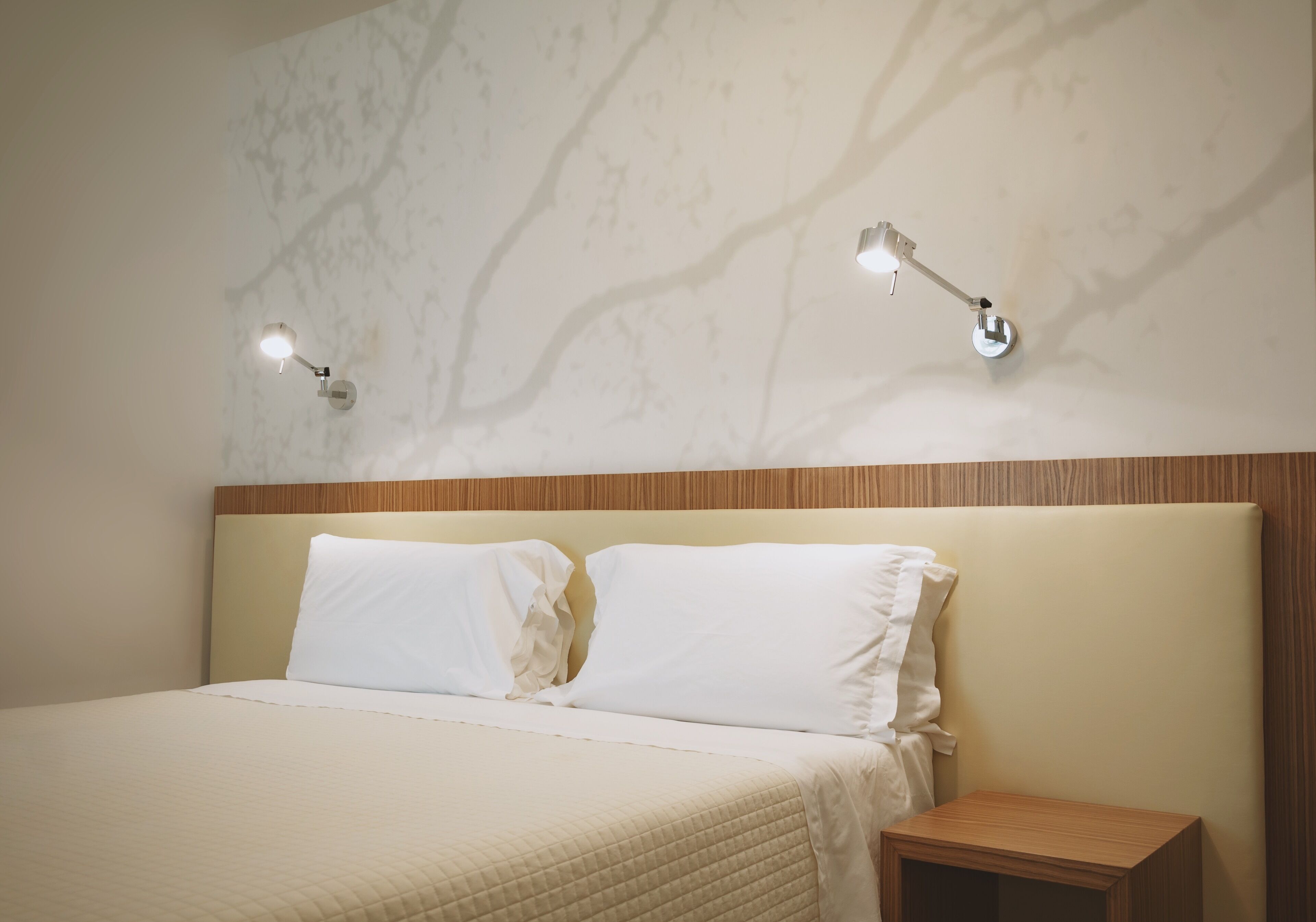A neatly made bed with white linens, a wooden headboard, and two wall-mounted reading lamps.