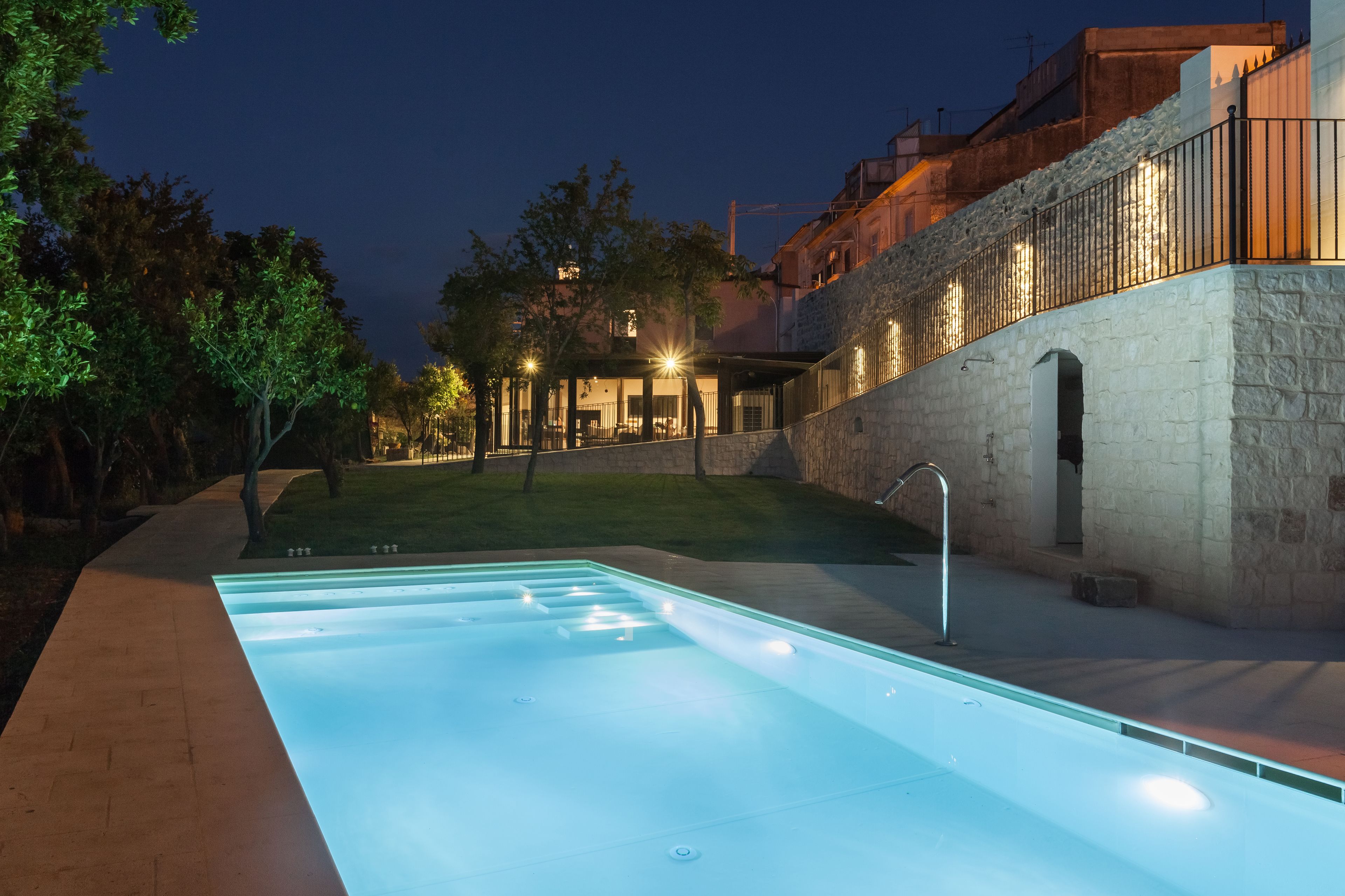 A swimming pool with a stone wall and a metal railing, surrounded by trees and a building with illuminated windows.