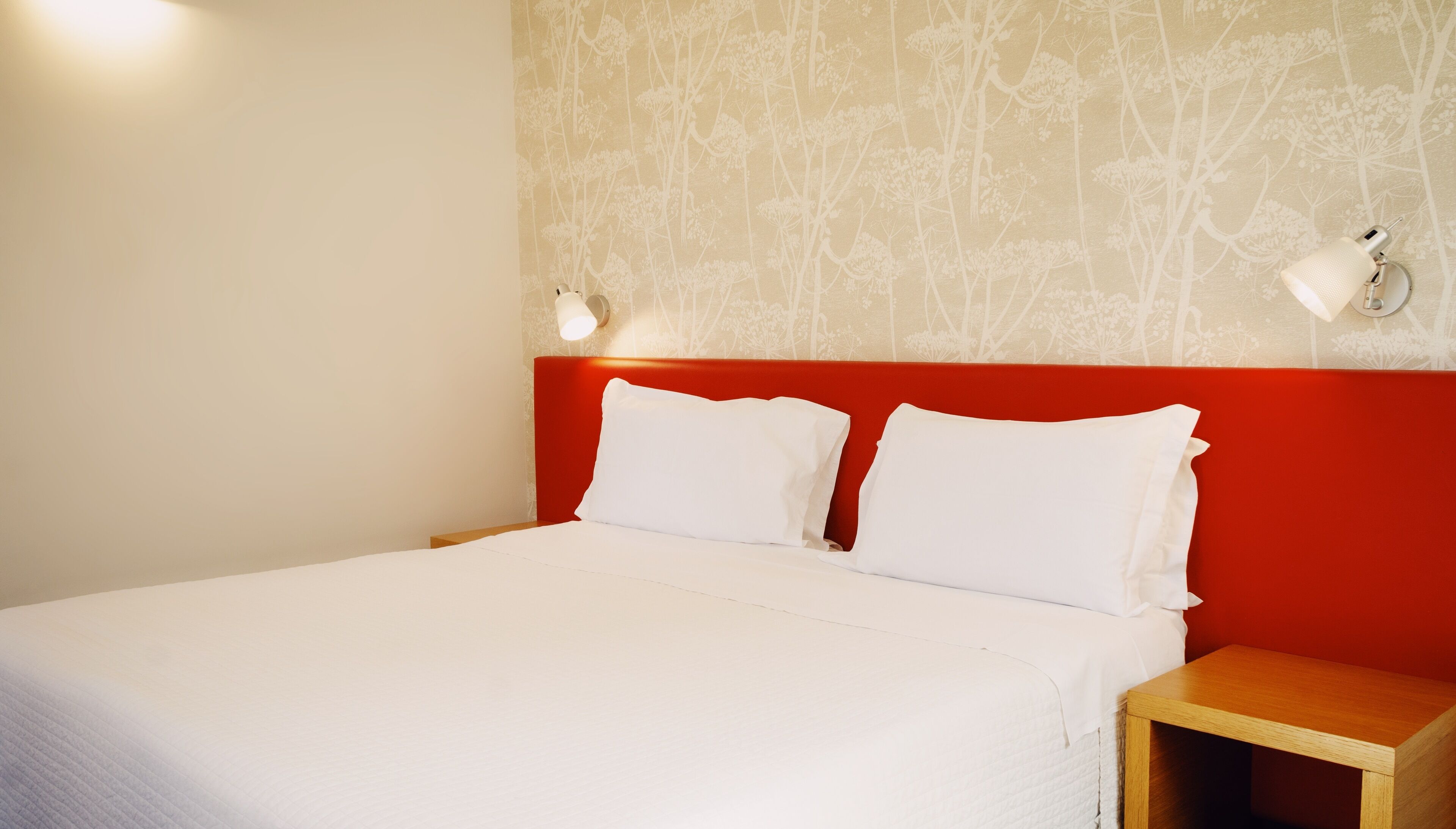 A hotel room with a bed, two bedside lamps, a red headboard, and a wooden nightstand.