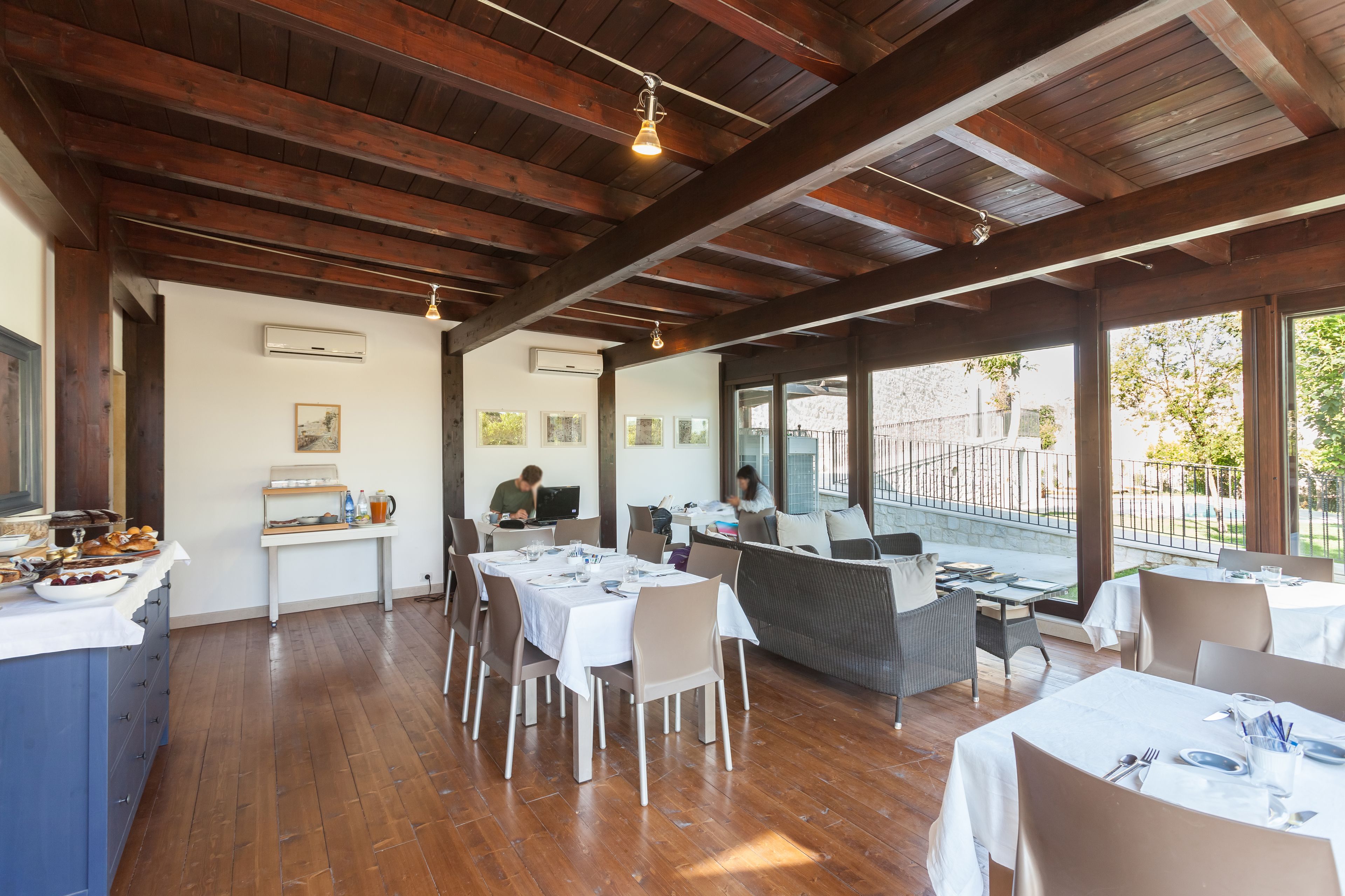 Un restaurante con vigas de madera, mesas y sillas blancas, y grandes ventanales con vistas a un jardín.