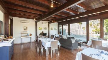 Un restaurante con vigas de madera, mesas y sillas blancas, y grandes ventanales con vistas a un jardín.