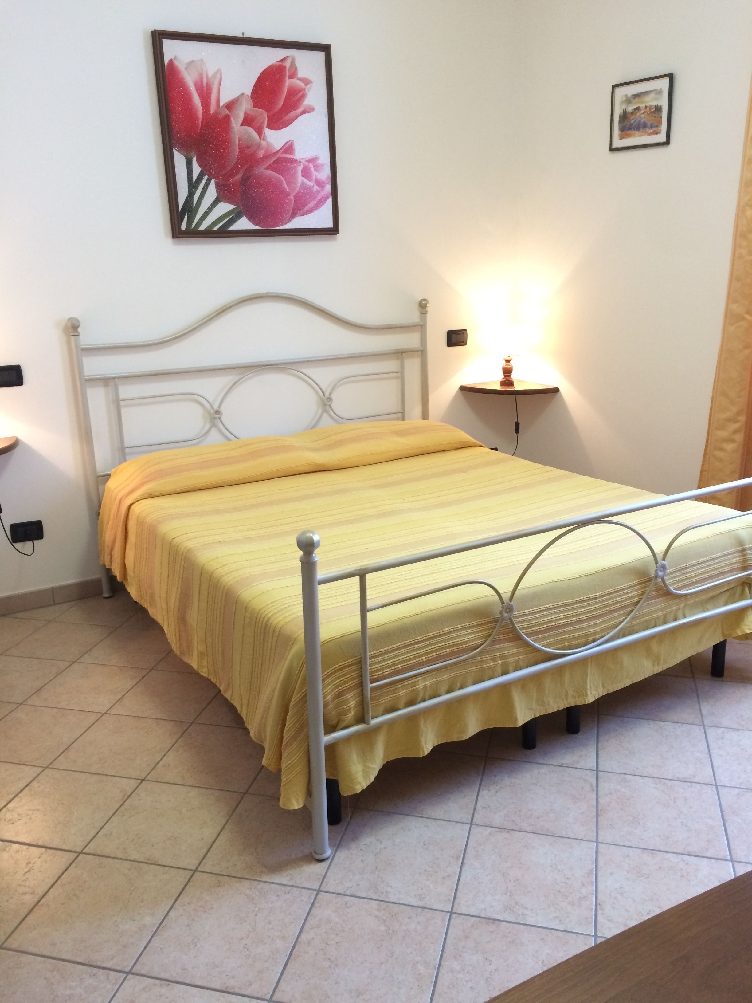 A bed with a metal frame and a yellow bedspread, a wall-mounted picture of flowers, a bedside table with a lamp, and a small picture frame on the wall.