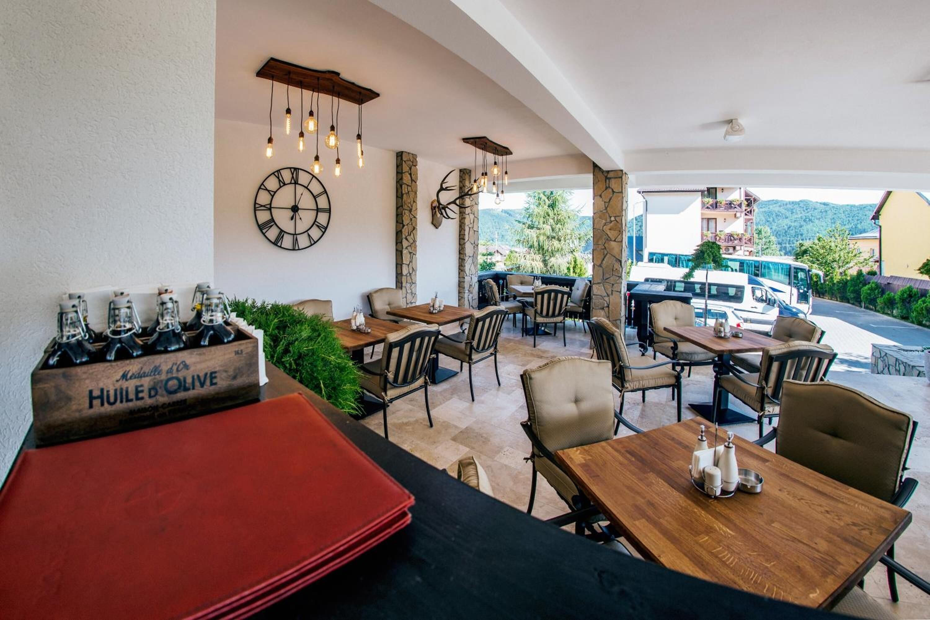 Een terras met stenen pilaren, houten tafels en stoelen. Er hangt een klok aan de muur en op een tafel staat een fles Huiles d'Olive.