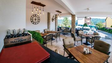 Een terras met stenen pilaren, houten tafels en stoelen. Er hangt een klok aan de muur en op een tafel staat een fles Huiles d'Olive.
