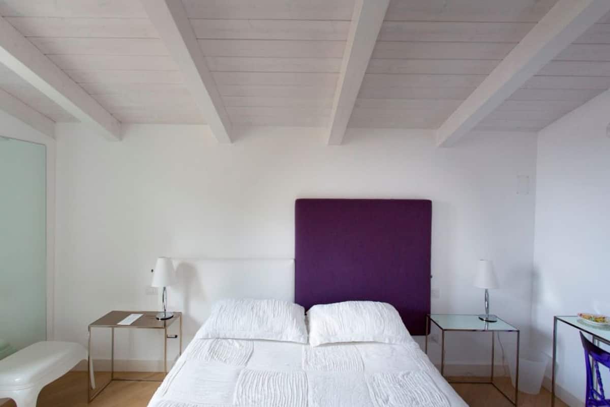 Ein Schlafzimmer mit einem Bett, zwei Nachttischen, einem violetten Kopfteil und einem kleinen Tisch mit einer Lampe.