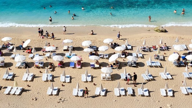 Une plage bordée de rangées de transats et de parasols, avec une eau d’un bleu limpide, où l’on peut voir des gens se baigner et se promener le long du rivage.