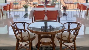 Un coin repas avec une table en verre et des chaises en osier, offrant une vue sur la côte, avec des bâtiments et des montagnes en toile de fond.