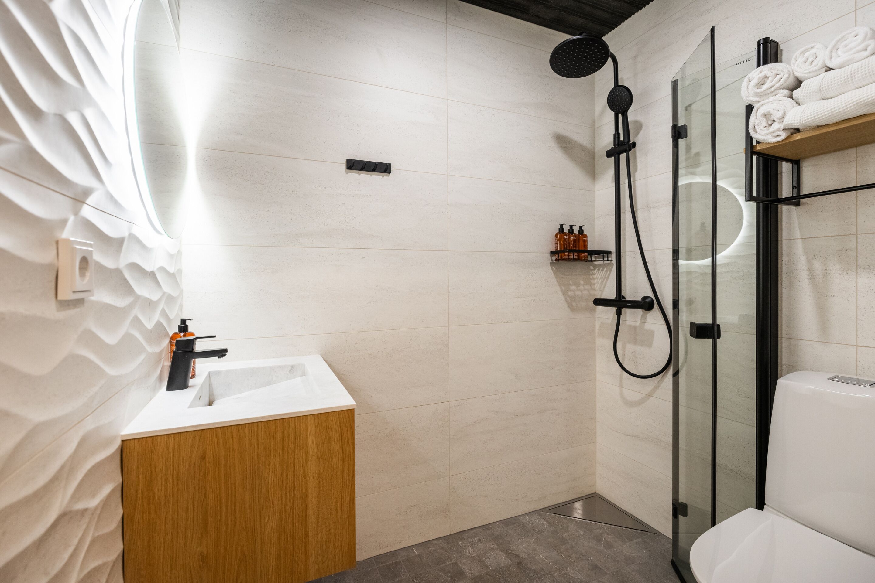 A modern bathroom with a wooden vanity, a glass shower enclosure, and neatly rolled towels on a shelf.