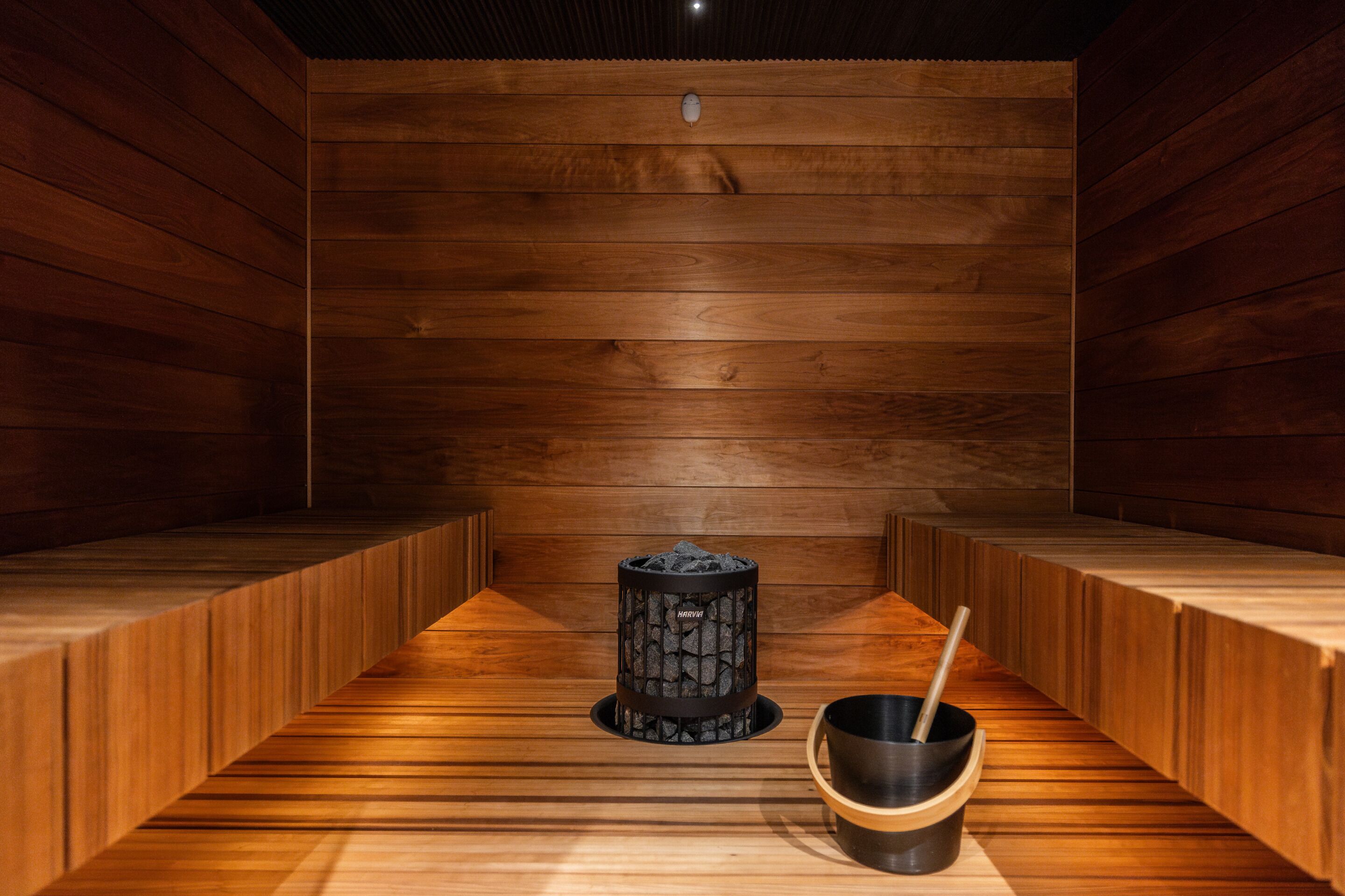 Un sauna avec des murs et des bancs en bois, un appareil de chauffage central et un seau avec une brosse.