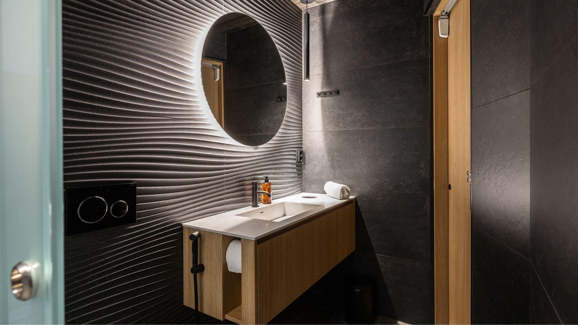 Un bagno moderno con un mobile contenitore in legno, uno specchio rotondo e una parete strutturata.