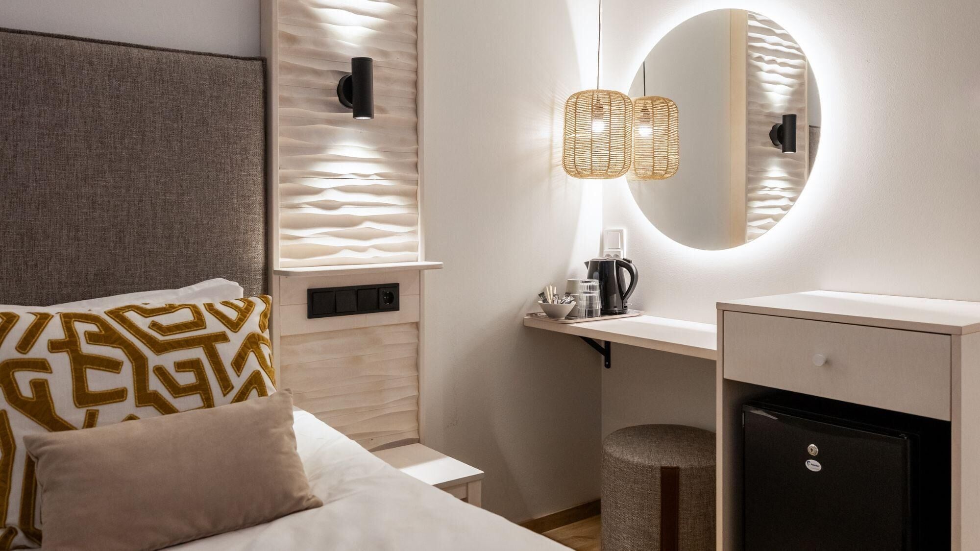 A hotel room with a bed, bedside table, a small desk with a chair, a mirror, and a wall-mounted light fixture.