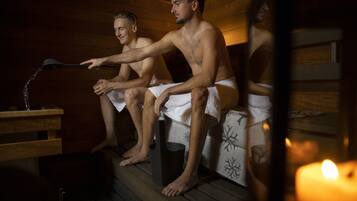 Two individuals in a sauna with wooden benches, towels, and a candle.