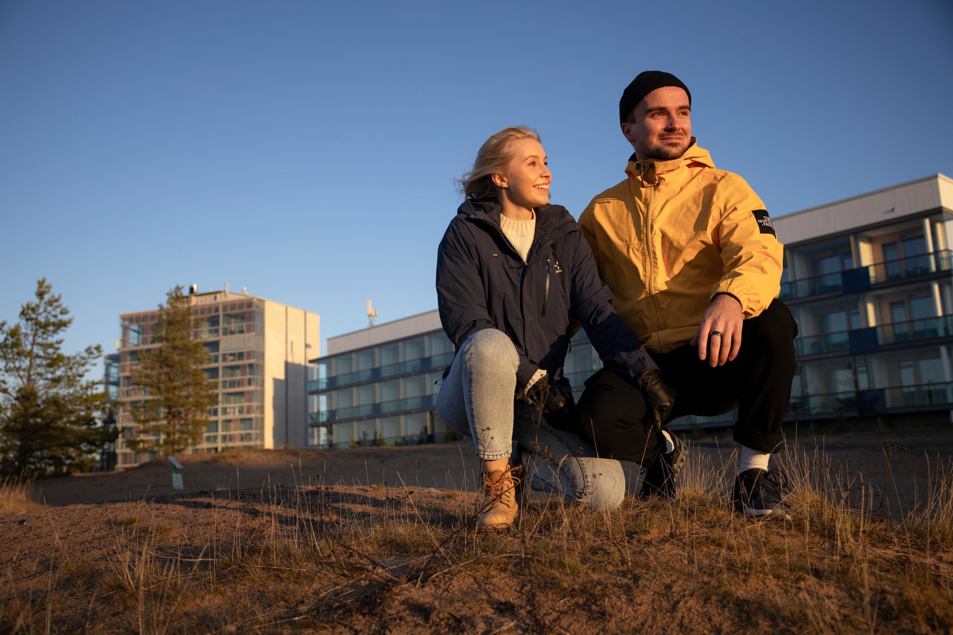 Dos personas sentadas en un césped, con un edificio moderno al fondo.