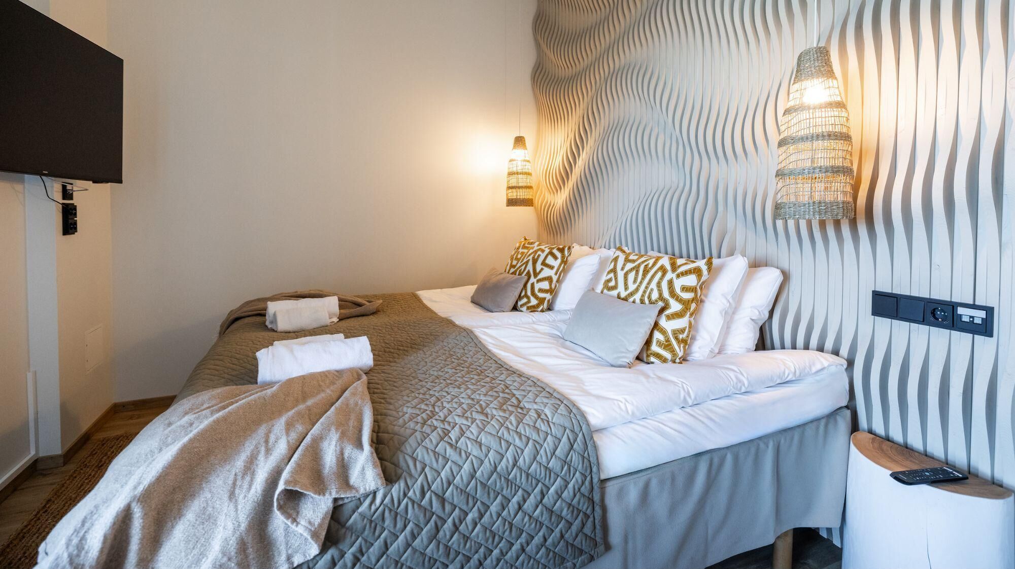 A neatly made bed with a quilted cover, a bedside table with a lamp, and a wall-mounted light fixture.