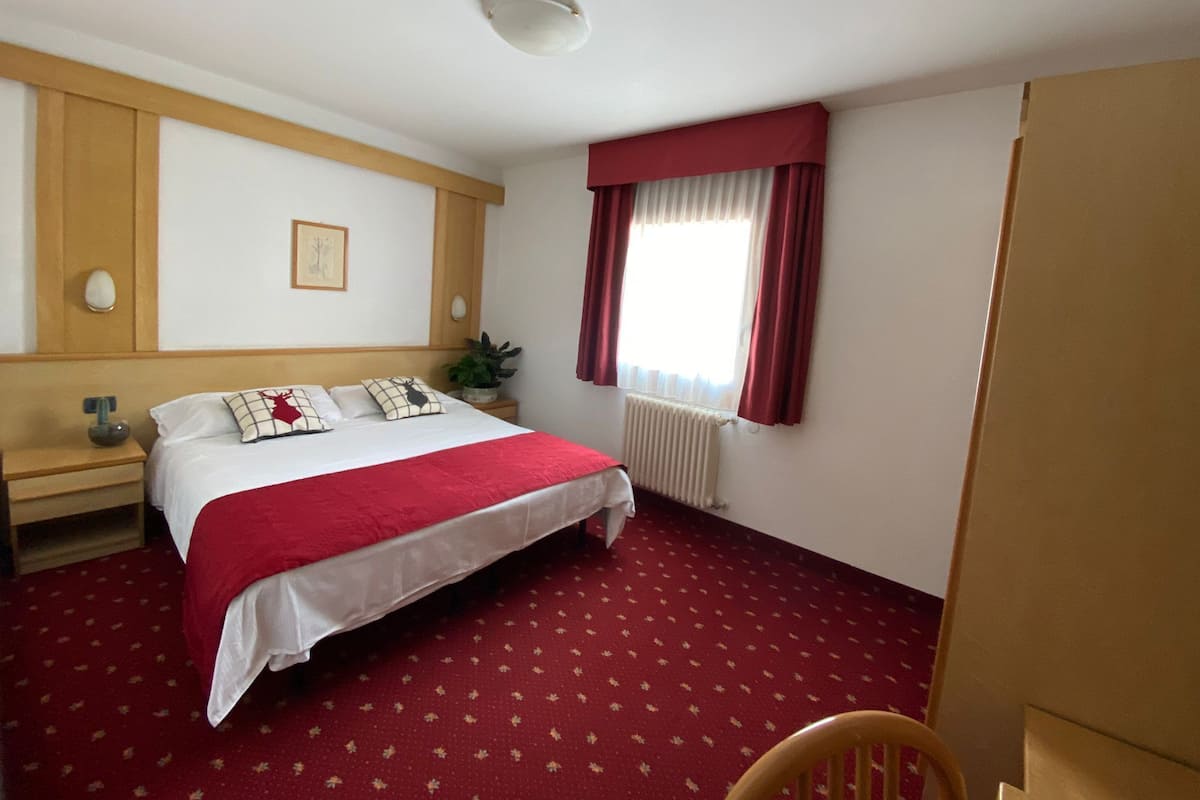 Habitación de hotel con una cama grande, cabecera de madera, alfombra roja, ventana con cortinas rojas y un radiador.