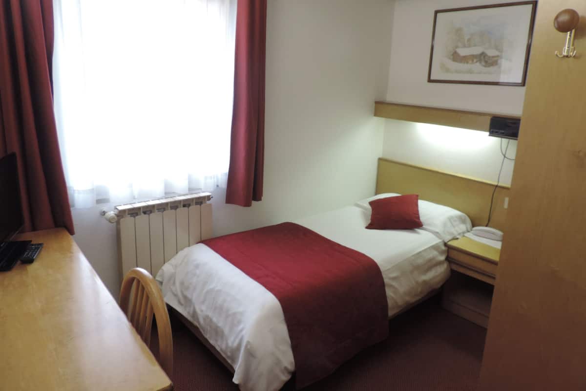 Habitación de hotel con cama, escritorio, silla, radiador, ventana con cortinas, mesita de noche, lámpara y un cuadro en la pared.