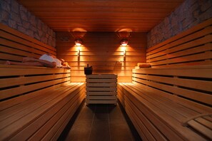 A sauna with wooden benches and stone walls, two wall-mounted lights, and a stack of towels in the center. - Grand Hotel Miramonti (Vermiglio)