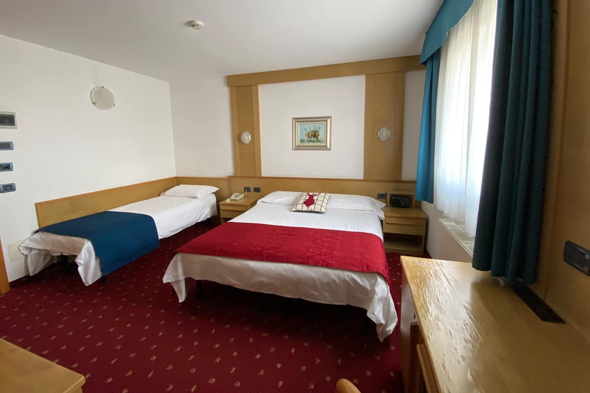 Habitación de hotel con dos camas individuales, cabecero de madera, alfombra roja, mesita de noche pequeña, teléfono, un reloj de pared y una ventana con cortinas.