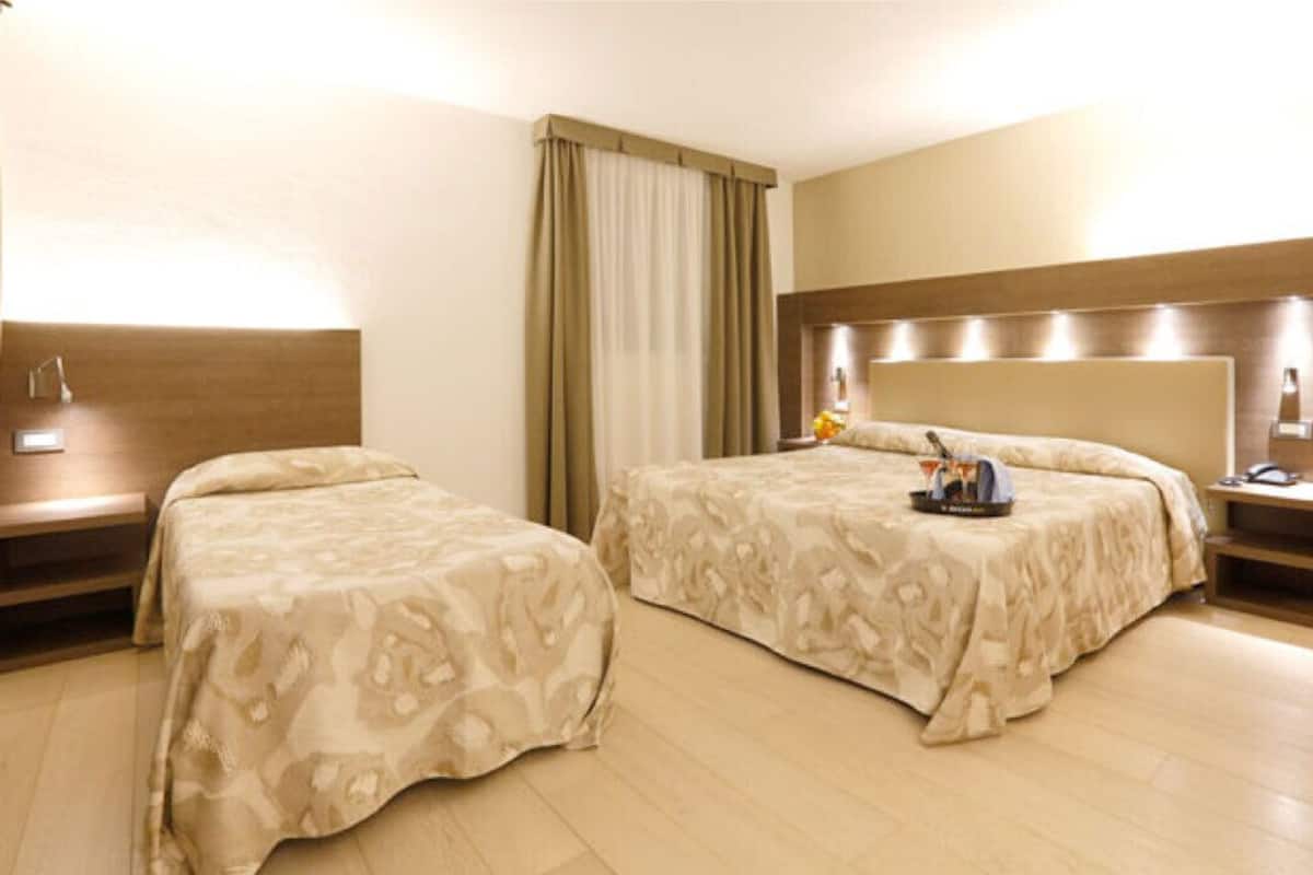 Una habitación de hotel con dos camas, cabecero de madera, mesitas de noche con lámparas y un gran ventanal con cortinas.