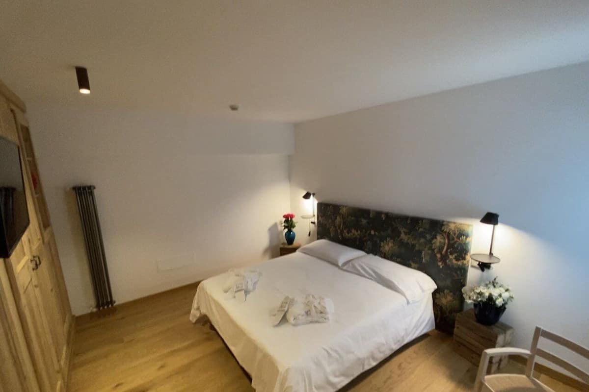 Una cama bien hecha con sábanas y toallas blancas, piso de madera, cabecera con estampado floral, mesita de noche con lámpara y una silla.