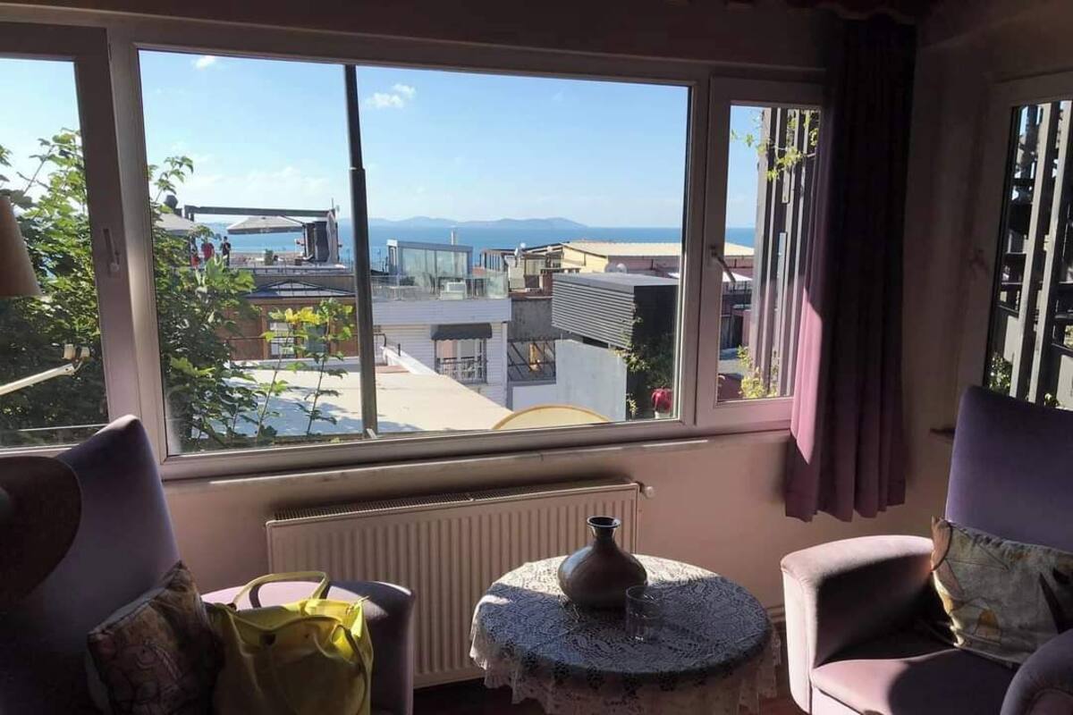 Ein Zimmer mit großem Fenster und Blick auf Gebäude und Berge. Ein Tisch mit einer Vase und einem Glas sowie zwei Stühle mit Kissen.