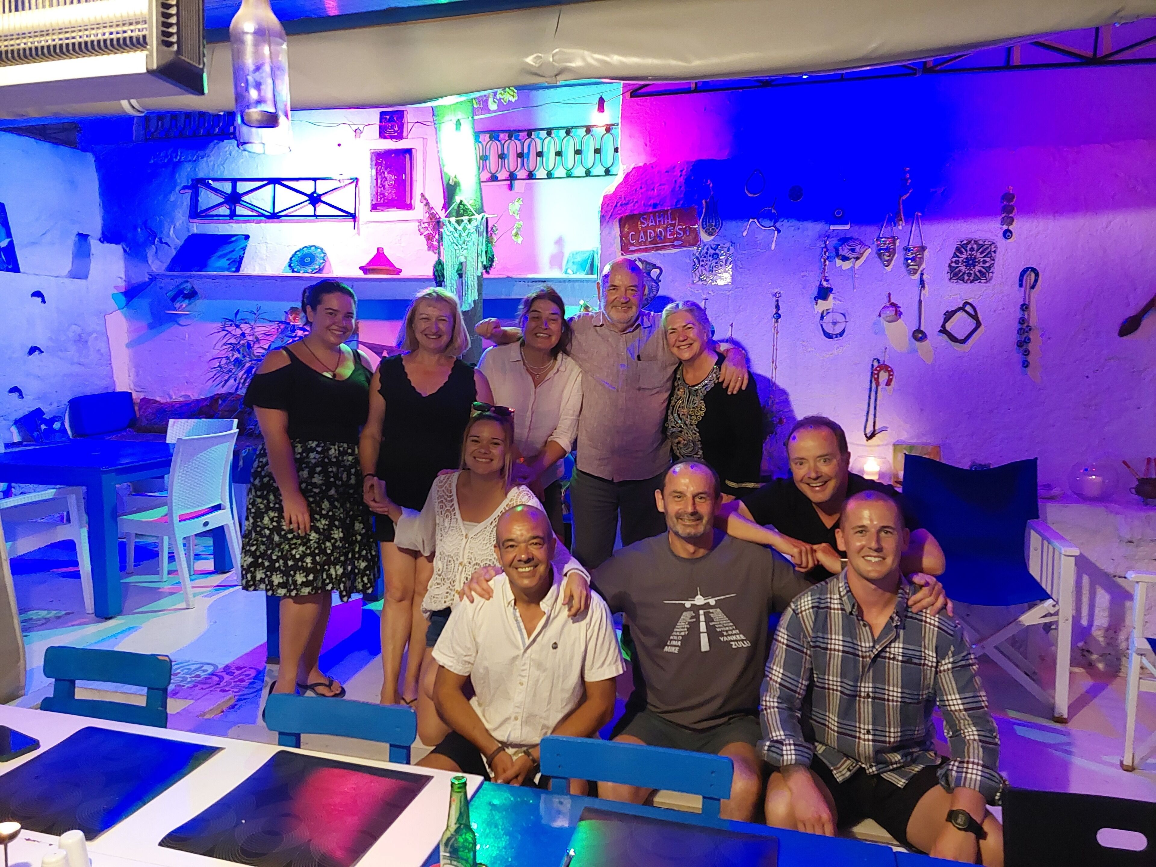 A group of people in a room with a table and chairs, a wall with hanging decorations, and a sign that reads "SAHIL CAPODEST".