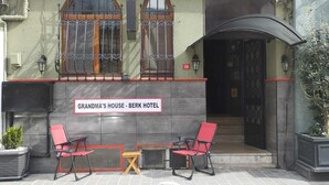 Exterior da Casa da Vovó - Berk Hotel, com uma placa, duas cadeiras vermelhas, uma pequena mesa e uma entrada curva.