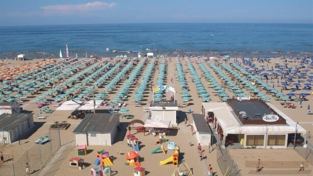 A beach with rows of umbrellas and beach chairs, a playground, and a building with a sign. - Hotel Mizar (Rimini)