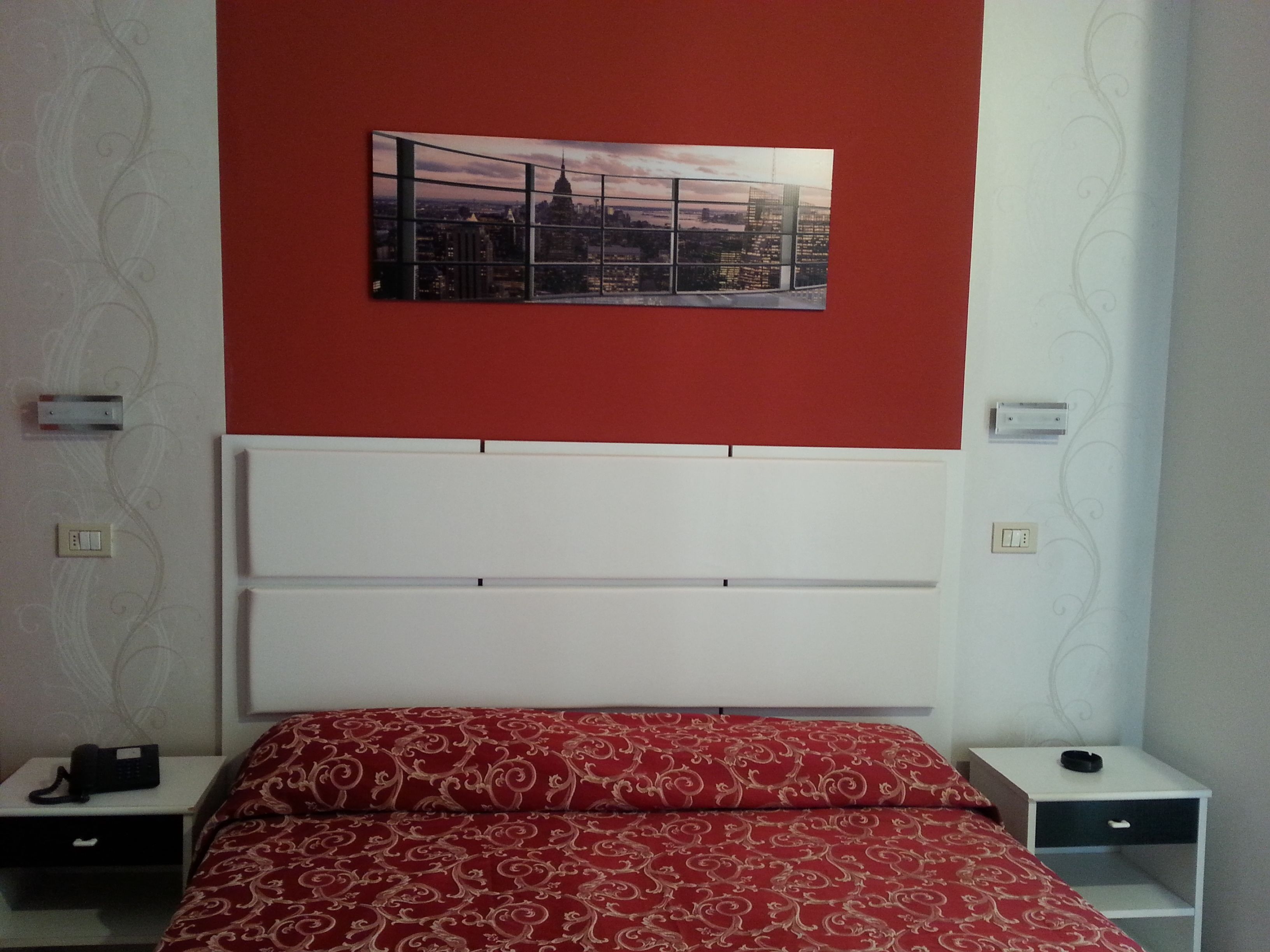 A bedroom with a red wall, a cityscape painting, a white headboard, and two bedside tables with lamps.