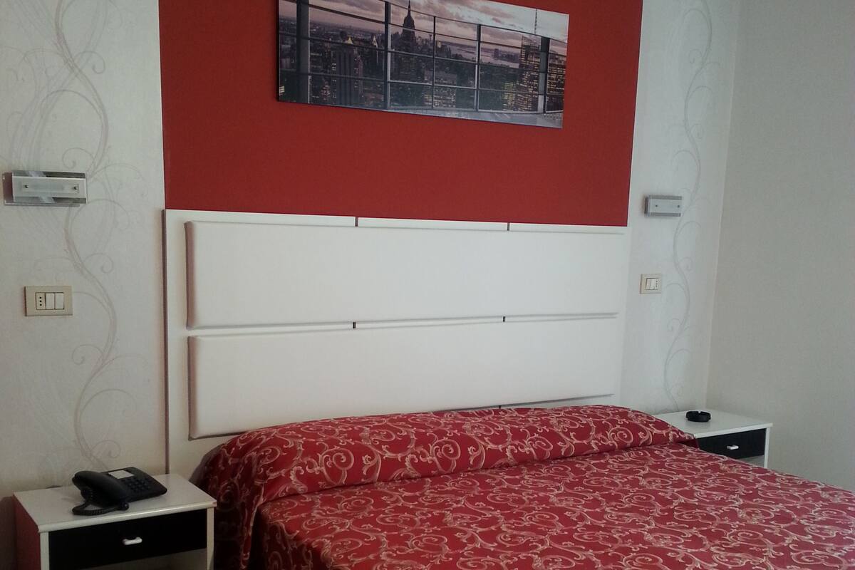 A hotel room with a red patterned bedspread, a white headboard, a small bedside table with a phone, and a wall-mounted picture of a cityscape.