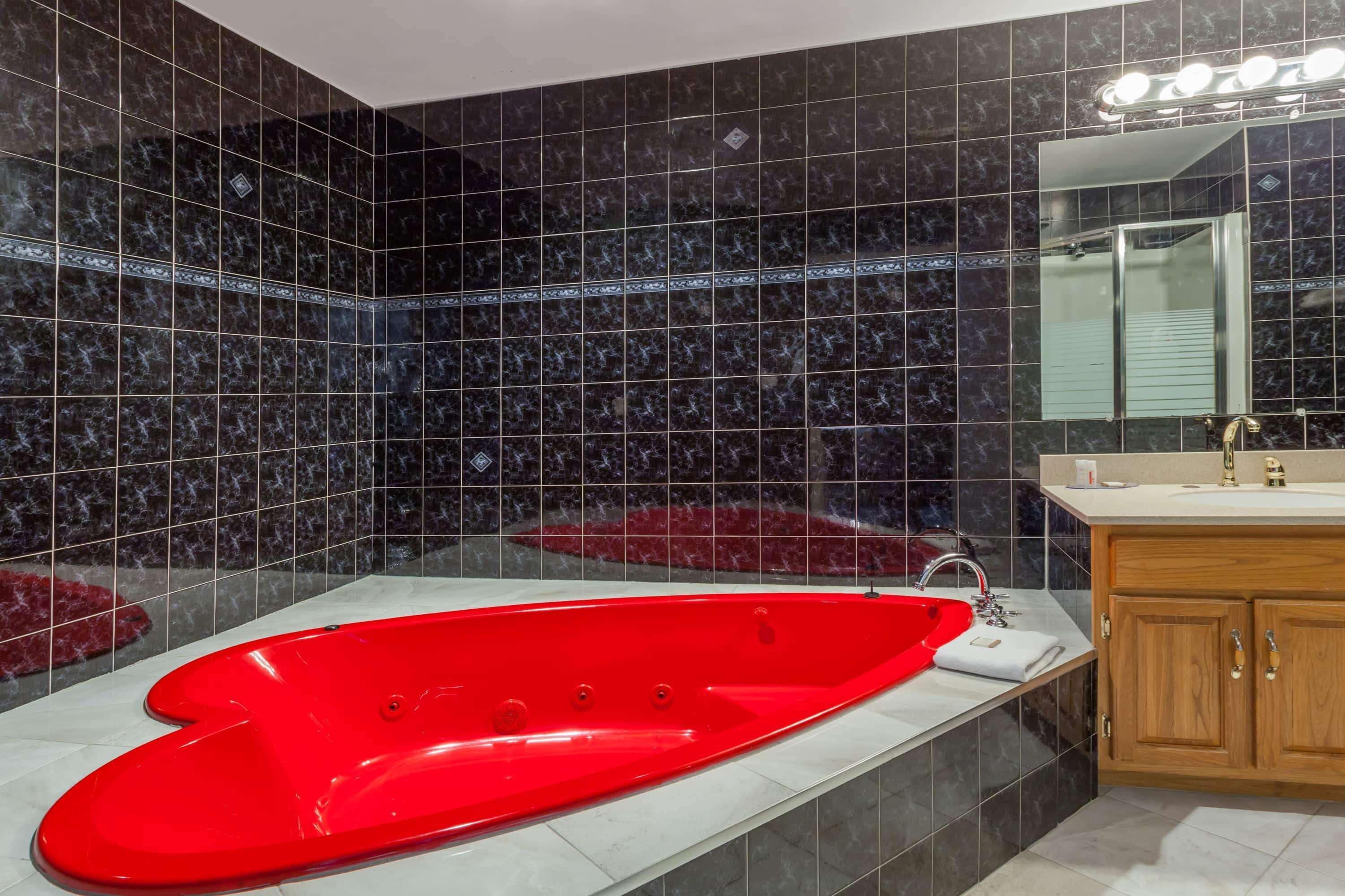 A bathroom with a red bathtub, dark tiled walls, a wooden vanity, and a mirror above the sink.