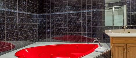 A bathroom with a red bathtub, dark tiled walls, a wooden vanity, and a mirror above the sink.