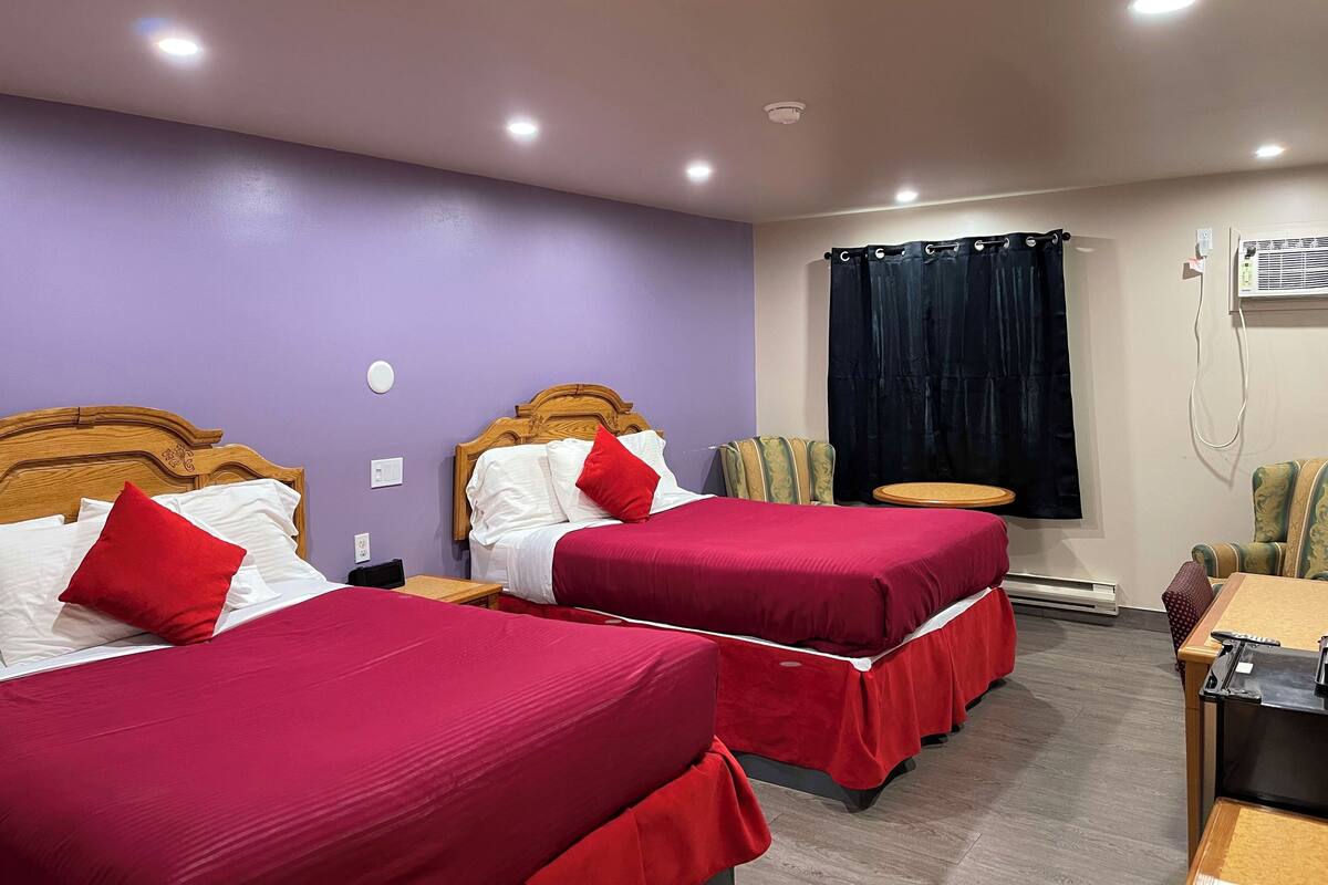 Habitación de hotel con dos camas, cada una con ropa de cama roja y cabeceros de madera. Hay una mesita y una silla, una ventana con cortina oscura y un equipo de aire acondicionado en la pared.