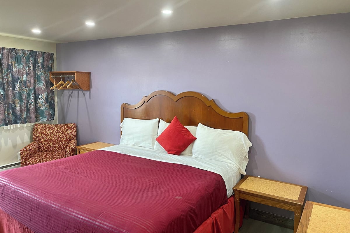 Un dormitorio ordenado con una cama de madera, una colcha roja y blanca, una almohada roja y un sillón con estampado.
