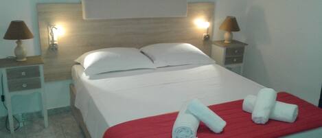 Uma cama bem arrumada com lençóis brancos e um cobertor vermelho, duas luminárias de cabeceira e um quadro na parede.