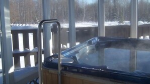 A hot tub with a wooden deck and metal railings, surrounded by a glass enclosure with a view of a snowy landscape and trees. - North Adventure Inn (Cochrane)