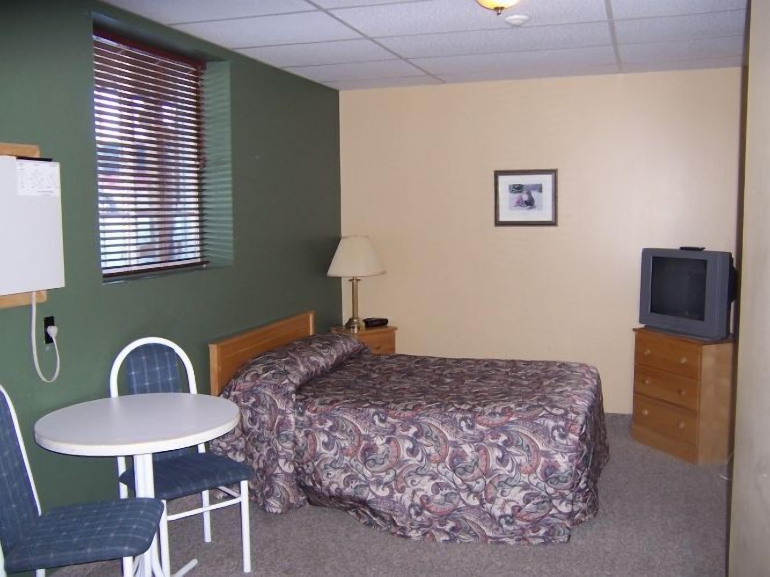 Una habitación de hotel pequeña y sencilla, con cama, una silla, una mesita, televisión y ventana con persianas.