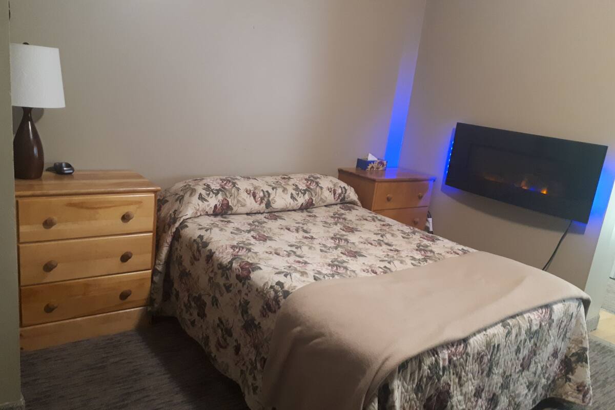 Une chambre à coucher comprenant un lit, une commode en bois, une table de chevet, une lampe et un téléviseur fixé au mur.