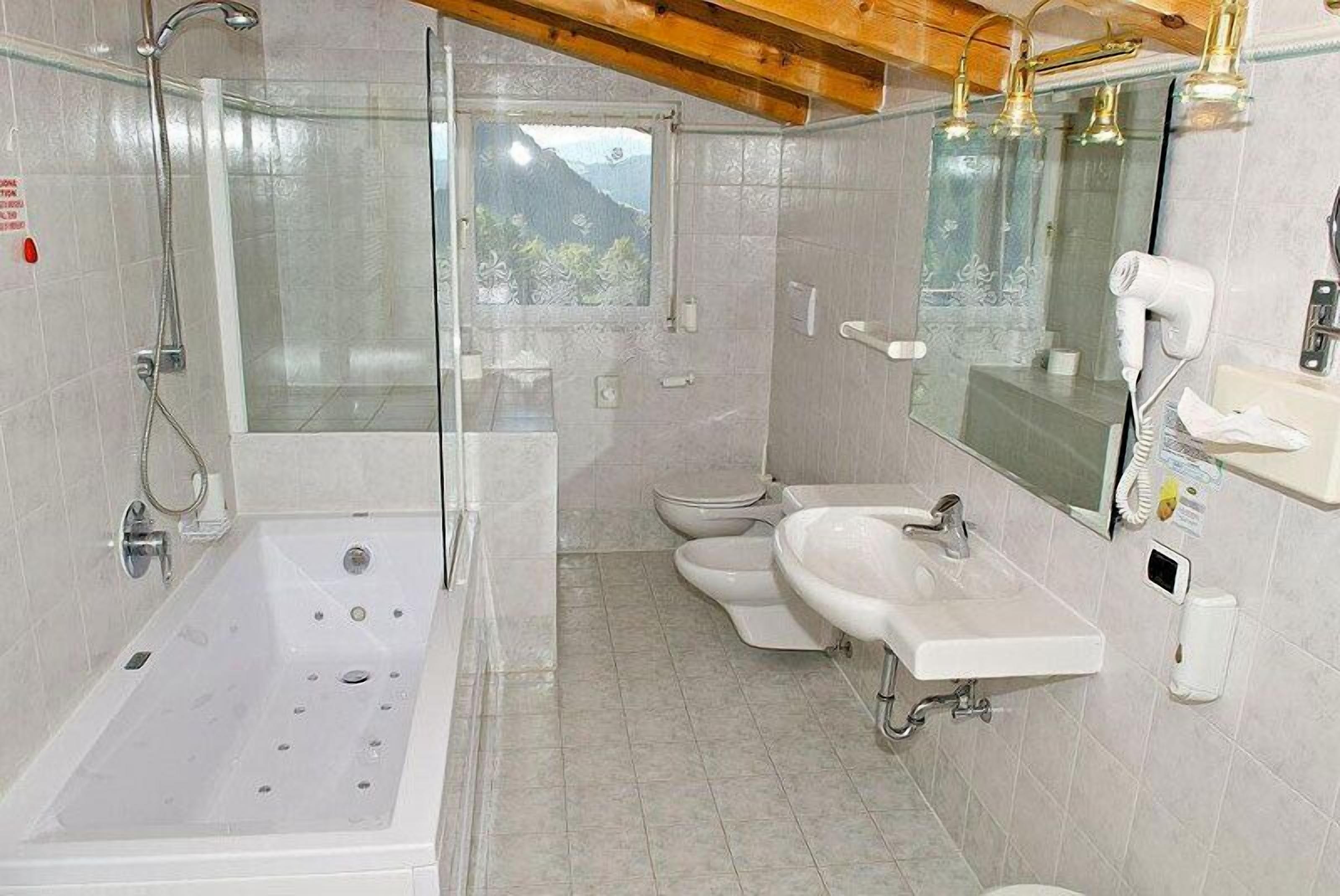 Un baño moderno con ducha de mampara de vidrio, una bañera grande, dos inodoros, un lavabo y un espejo.
