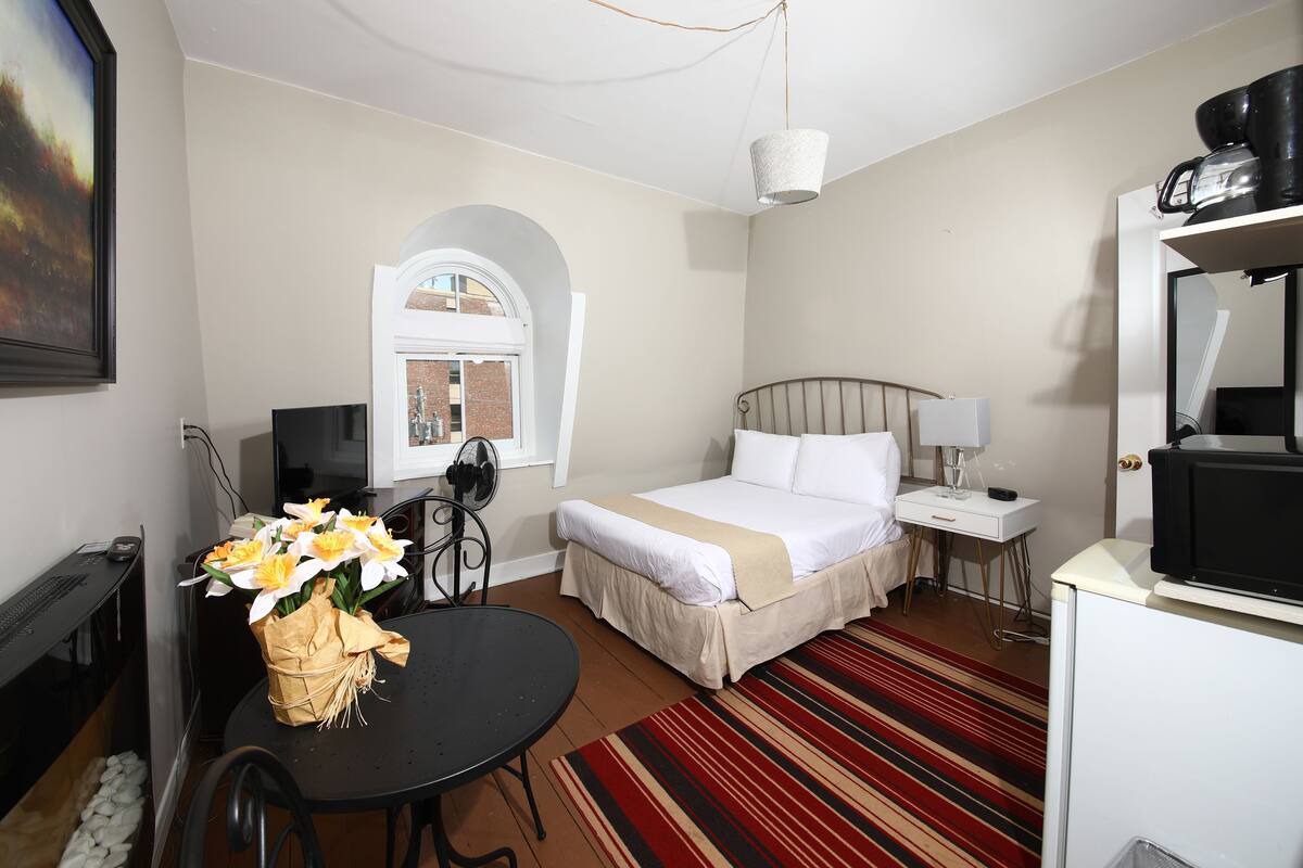 A hotel room with a bed, bedside table, lamp, TV, desk, chair, and a bouquet of flowers.