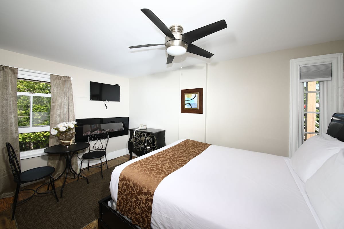 A hotel room with a bed, a desk with a chair, a TV, a ceiling fan, and a window with curtains.