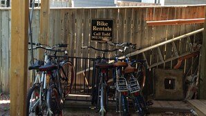 Una estación de alquiler de bicicletas de madera, con un letrero anunciando el alquiler, suelo de ladrillo y una valla de madera al fondo.