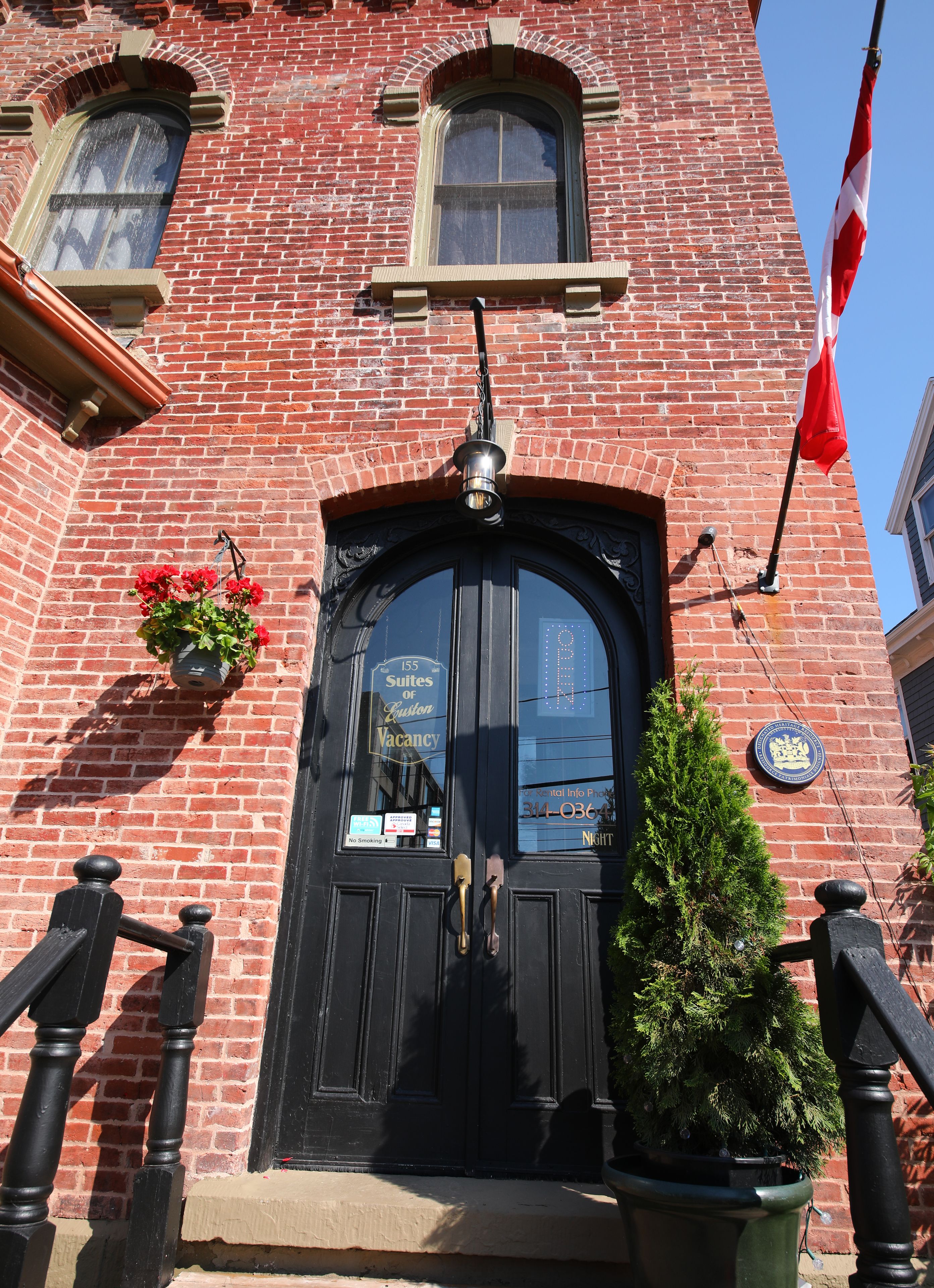 A red brick building with a black door, a window with a hanging flower pot, and a flag.