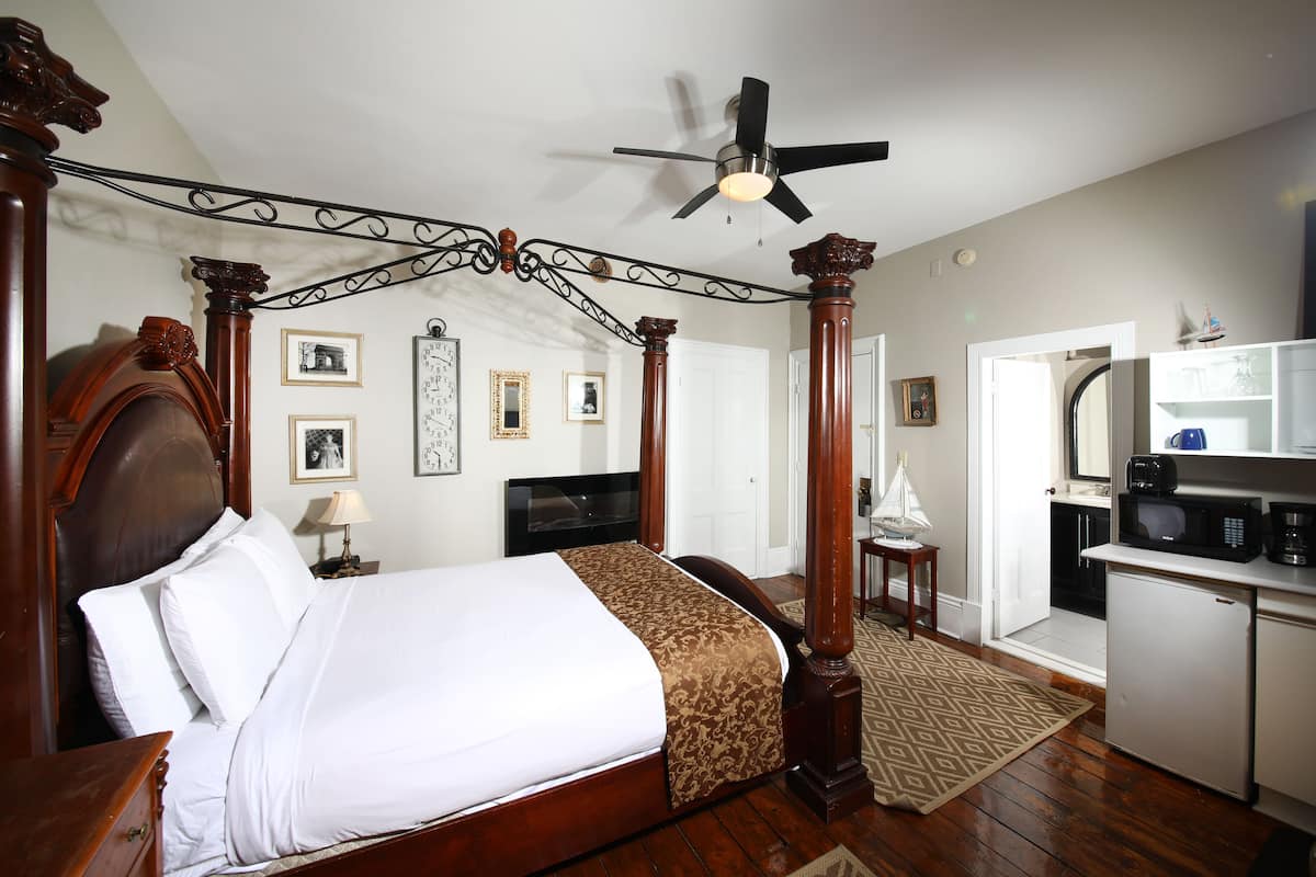A four-poster bed with a ceiling fan, a small table with a lamp, and a kitchen area with a refrigerator and microwave.