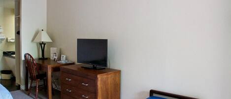 A hotel room with a bed, a desk with a TV, a dresser, a chair, and a small table with a lamp.