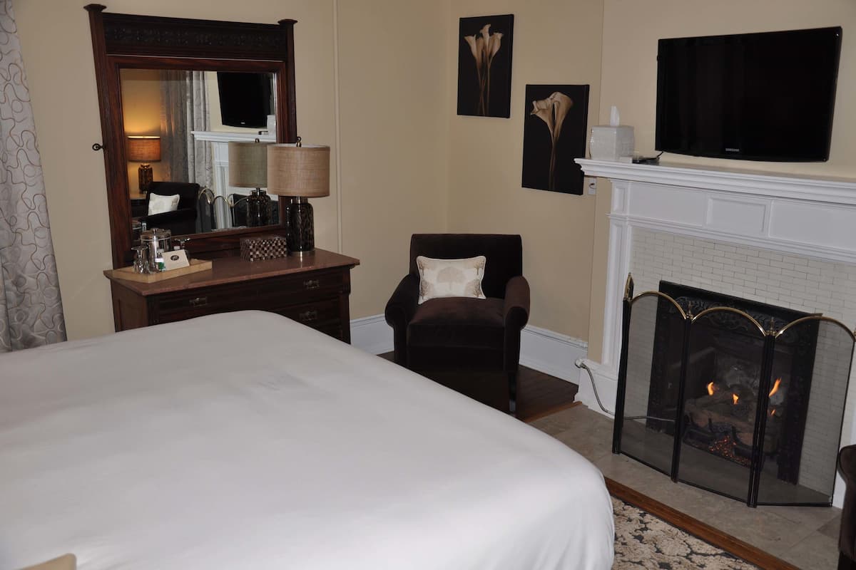 A hotel room with a large bed, a wooden dresser, a chair, a fireplace, and a television mounted above the fireplace.