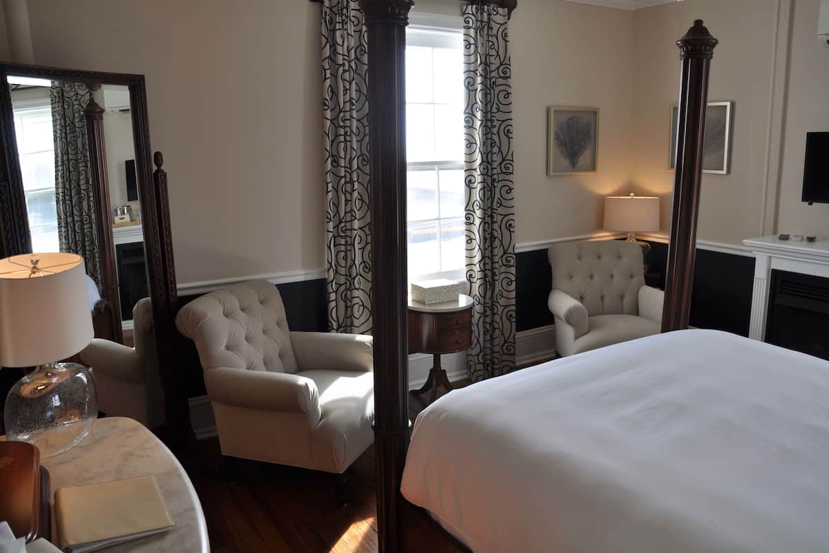 A hotel room with a large bed, two armchairs, a small table with a lamp, and a mirror.